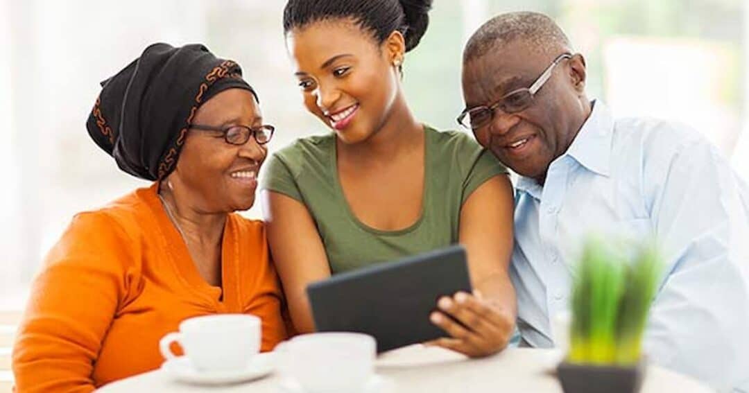A young woman sits between an elderly woman in an orange cardigan and an elderly man in glasses. They are looking at a tablet while sitting at a table with cups and a small plant. All are smiling and appear engaged with the device.