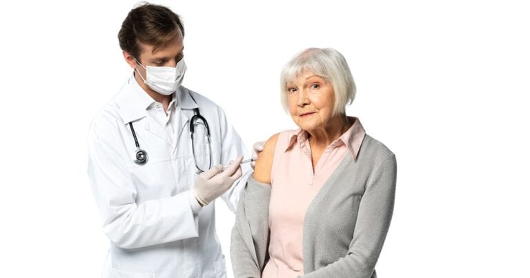 A doctor in a white coat and mask is administering a vaccine to an elderly woman with short white hair, wearing a pink shirt and gray cardigan. The background is plain white.