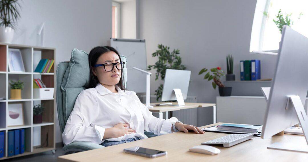 a woman sitting at her desk at work holds her stomach due to nausea