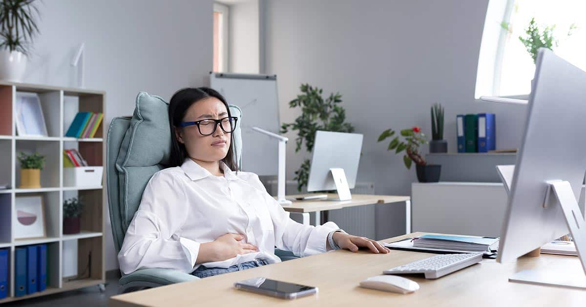 a woman sitting at her desk at work holds her stomach due to nausea