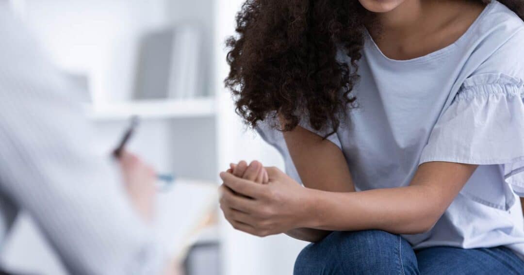 A person with curly hair is sitting, clasping their hands together. Another person in the foreground is holding a pen and notepad, suggesting a conversation or counseling session. The background is softly blurred.