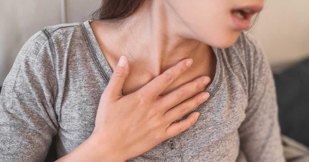 A person wearing a gray top is clutching their chest, seemingly in discomfort or experiencing difficulty breathing, possibly due to anaphylaxis. Their face is partly visible, and their posture suggests concern.