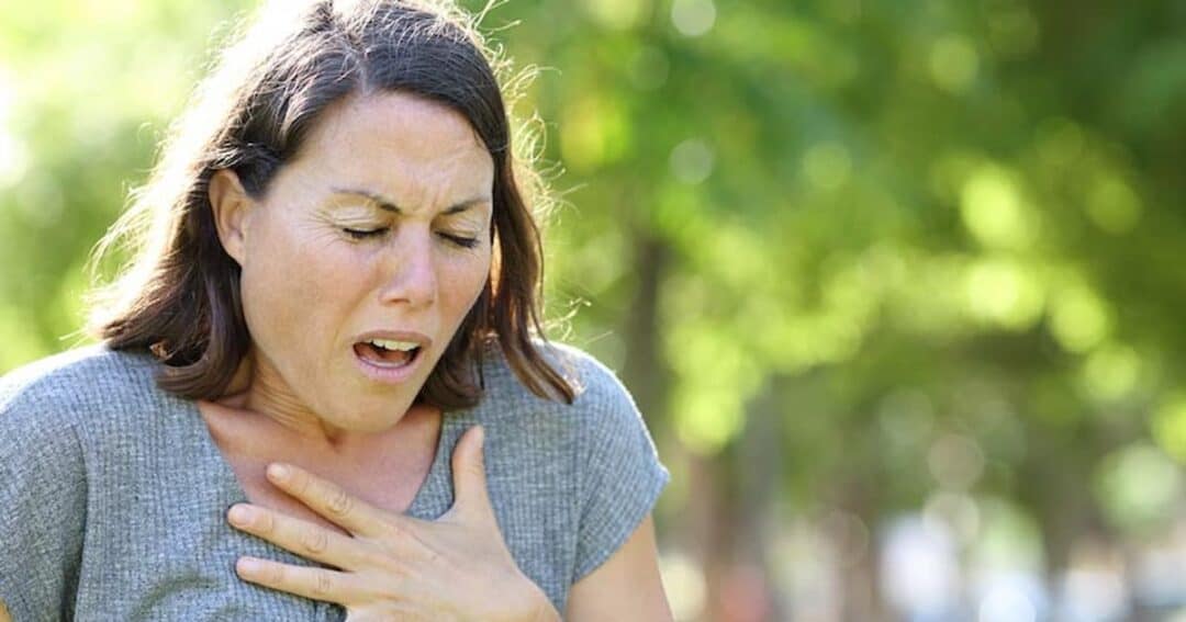 A woman outdoors appearing to have difficulty breathing, with her hand on her chest. She looks concerned, standing against a blurred background of green trees.