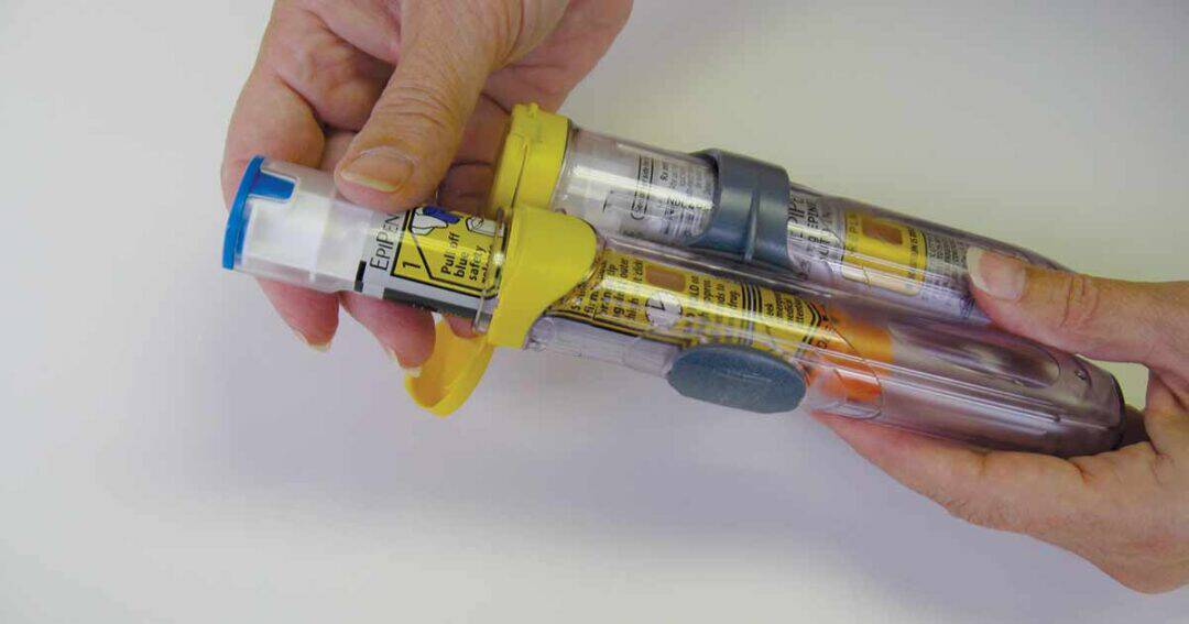 Two hands hold two epipens, one with a yellow cap and the other with an orange cap, against a white background. The auto-injectors have labels and instructions printed on them.