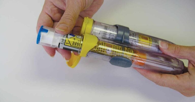 Two hands hold two epipens, one with a yellow cap and the other with an orange cap, against a white background. The auto-injectors have labels and instructions printed on them.