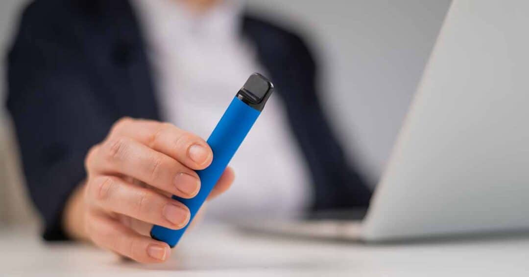 A person holds a blue vape pen in their hand, with a laptop in the background. The individual is wearing a dark blazer, and their focus seems to be on the vape pen.