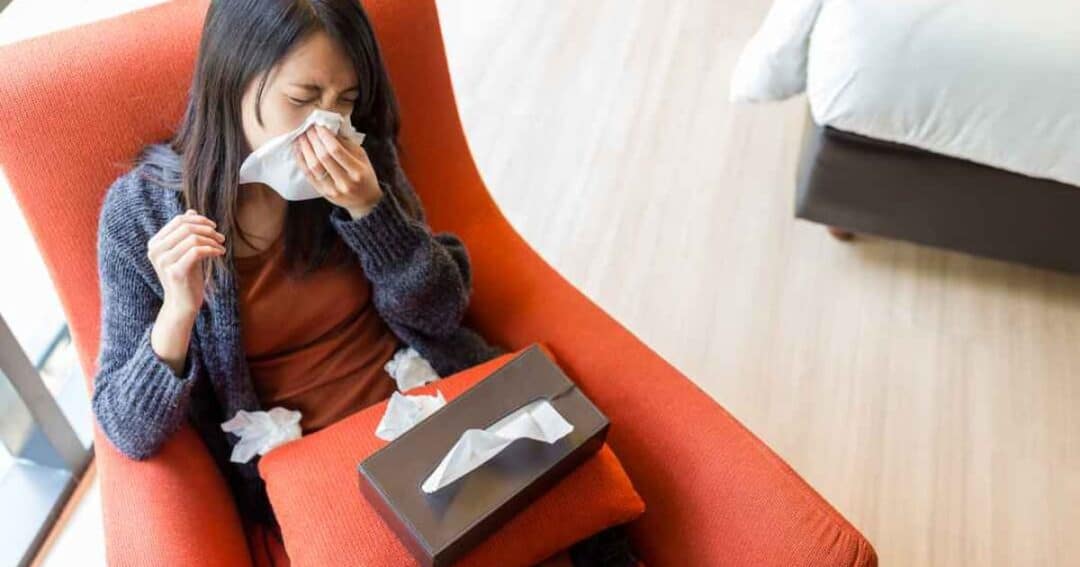 A woman sits on an orange armchair, holding a tissue to her nose. A tissue box rests on her lap, with used tissues scattered around. She appears unwell, possibly dealing with a cold or allergies. A bed is visible in the background.