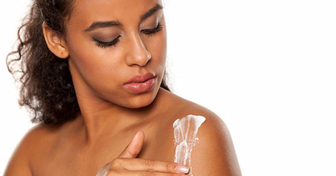 A woman applying lotion to her shoulder. She has curly hair and is looking down at the lotion with a neutral expression. The background is plain white.