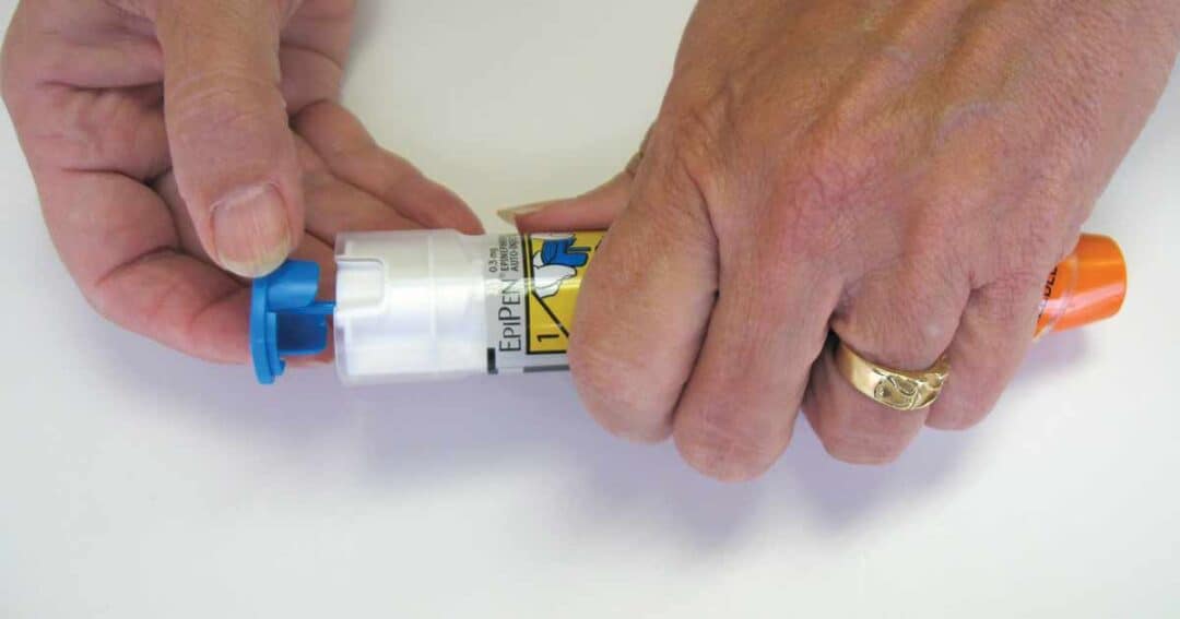 A person carefully holds an EpiPen, their hands moving deftly to remove the blue safety cap. The device features a distinct yellow label and an orange tip, while a ring subtly adorns their left finger, reflecting light.