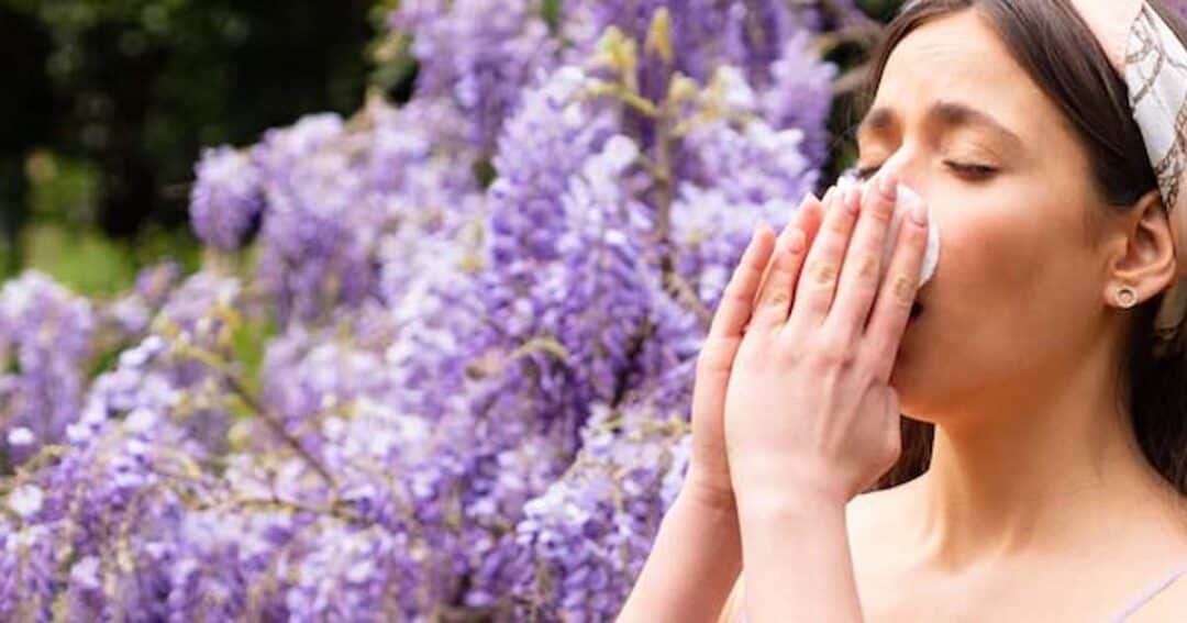 A woman with long brown hair stands in front of blooming purple flowers, holding a tissue to her nose with both hands, appearing to sneeze. She wears a light headband, and the background is filled with greenery.
