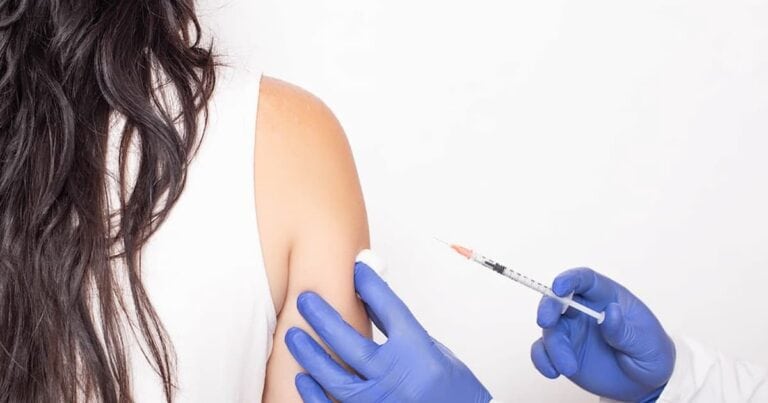 Person with long hair receiving a vaccine injection in their upper arm. A healthcare professional wearing blue gloves administers the shot. The background is plain and white.