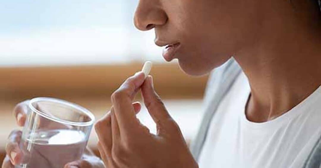 A person holds a pill in one hand and a glass of water in the other, preparing to take medication. The focus is on the person's face and hands. The background is softly blurred.
