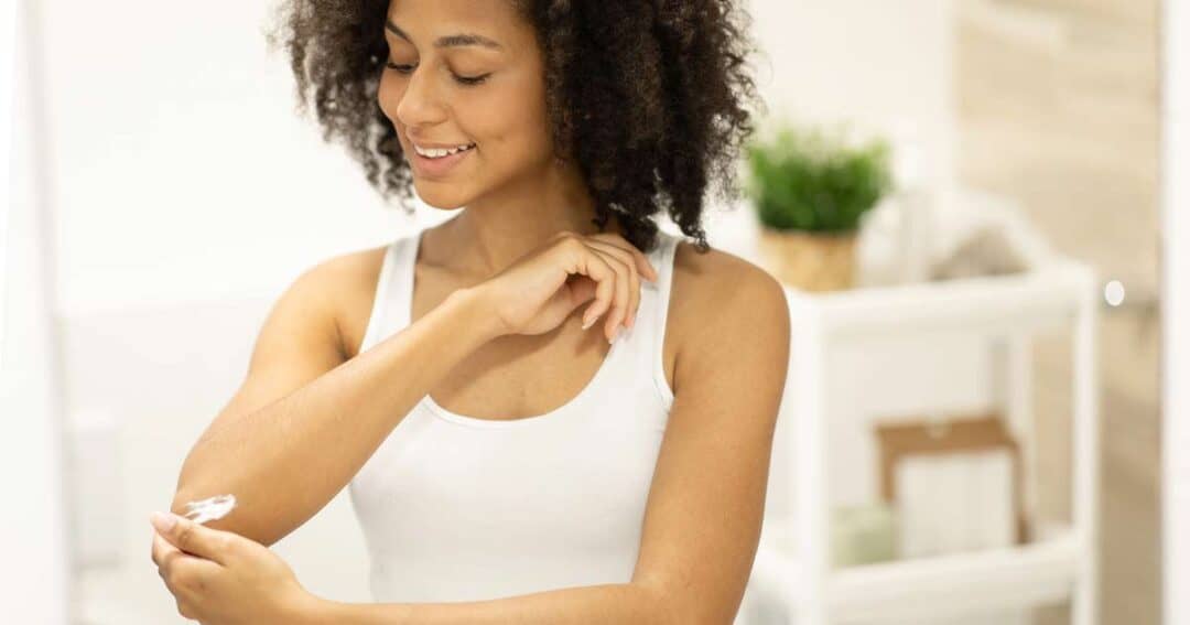 A person in a white tank top is applying eczema treatment lotion to their elbow in a bright bathroom. The background includes shelves with plants and towels. They are smiling and looking at their arm.