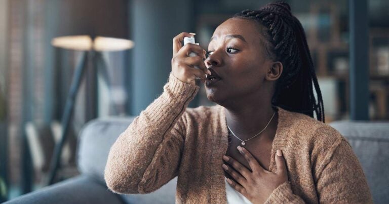 A person sitting on a couch uses an asthma inhaler. They have one hand on their chest and are wearing a brown sweater. The background features a lamp and a blurred interior setting.