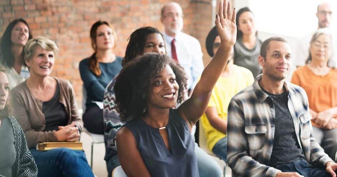 A diverse group of people is seated in a room with visible brick walls. A woman in the foreground is smiling and raising her hand. Others around her appear attentive and engaged.