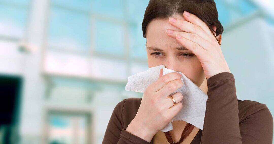 A woman holding a tissue to her nose and touching her forehead, appearing to have a cold or allergies, with a blurred indoor background.