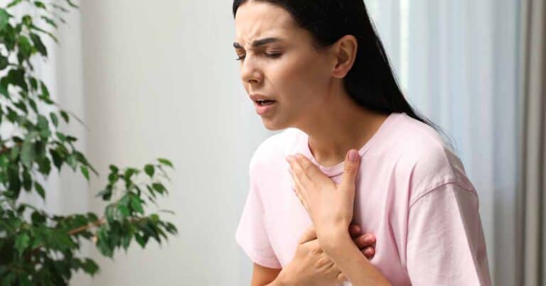A woman in a pink shirt is clutching her chest with both hands, appearing to be in pain or discomfort. She is indoors, with a leafy plant in the background and light coming from a window.