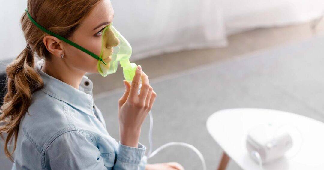 A woman with blonde hair wearing a green oxygen mask sits indoors, holding a nebulizer mouthpiece connected to a machine on a nearby white table. She looks to the side, appearing calm and focused.