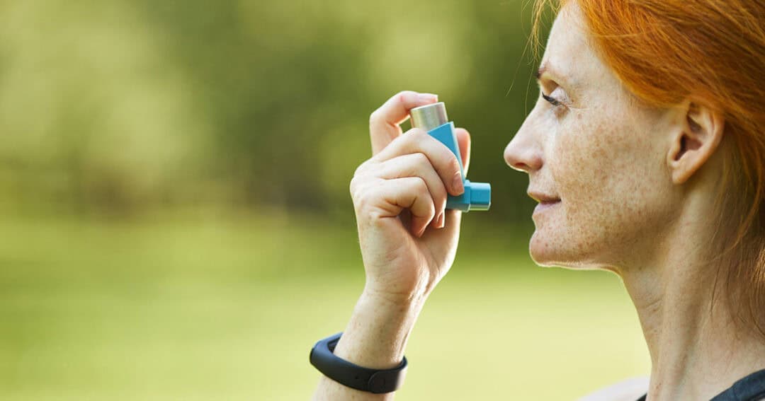 A woman with red hair is using her asthma inhaler outdoors. She holds it near her mouth, gazing into the distance against a blur of green in the background. A black wristband adorns her wrist, adding a subtle touch to the scene as she manages asthma relief with ease and poise.