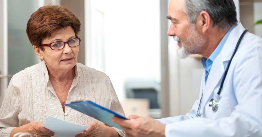 An elderly woman wearing glasses sits across from a male doctor in a white coat. The doctor is holding a clipboard and appears to be discussing medical information with her.