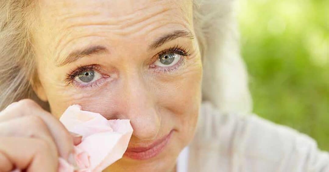 Grass Allergy 4 A woman with light-colored hair wipes her eye with a tissue while outdoors. She appears emotional or teary-eyed, with a background of greenery.