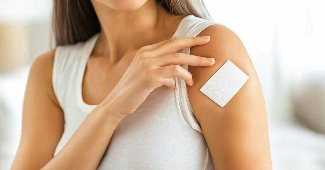A person wearing a sleeveless white top is touching a white adhesive patch on their upper arm. The focus is on the patch and the hand, with the background blurred.