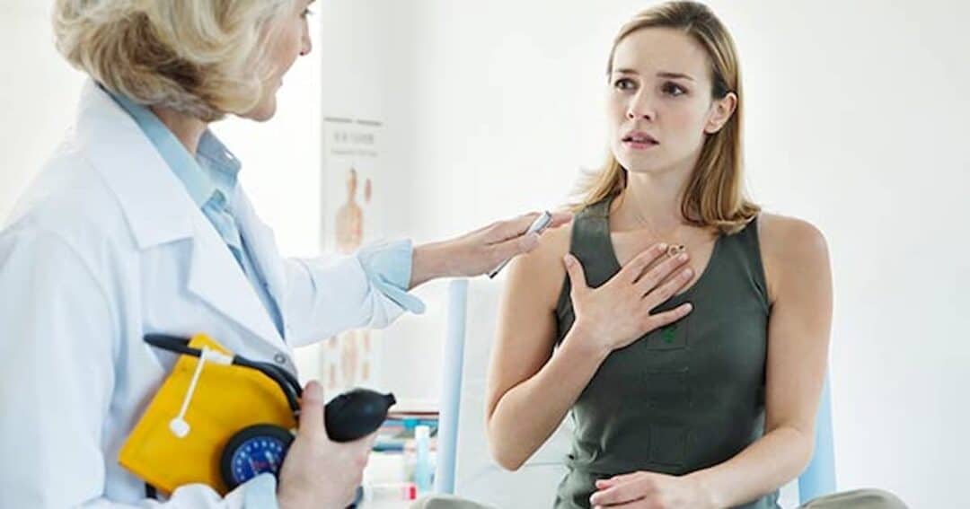 A doctor in a white coat is speaking with a concerned woman sitting on an examination table. The woman holds her hand to her chest, while the doctor holds medical equipment, possibly for testing or examination.