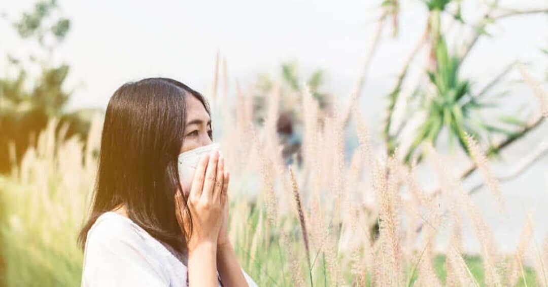 A woman with long hair stands outdoors, wearing a white mask over her nose and mouth. She is surrounded by tall grasses and trees, with sunlight filtering through the scene.