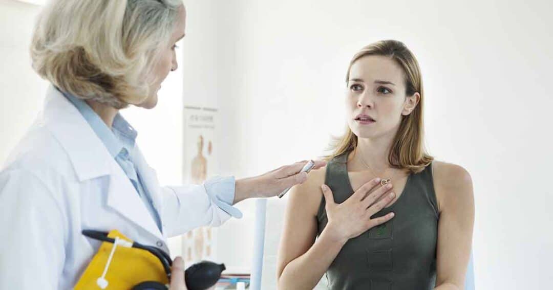 A doctor in a white coat gestures towards a woman who looks concerned, with her hand on her chest. They are in a medical office setting, and the doctor holds a blood pressure monitor.