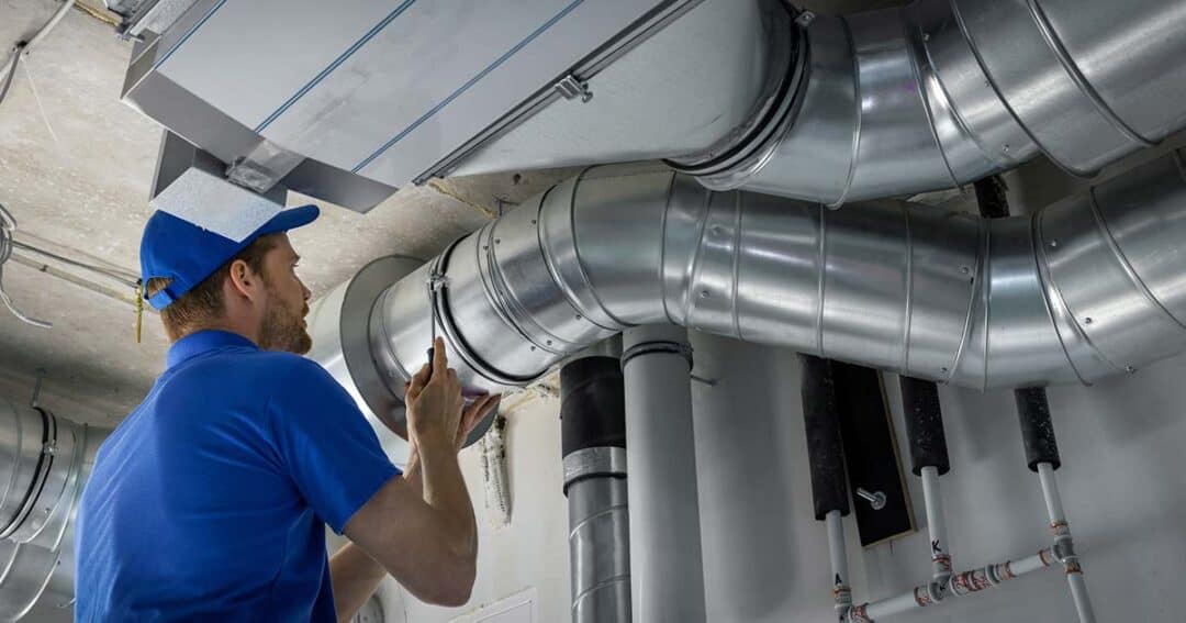Indoor Air Quality and Indoor Air Pollution 2 A maintenance worker in a blue uniform and cap inspects large metal air ducts overhead with a flashlight. The ducts are part of an industrial ventilation system in a utility room.