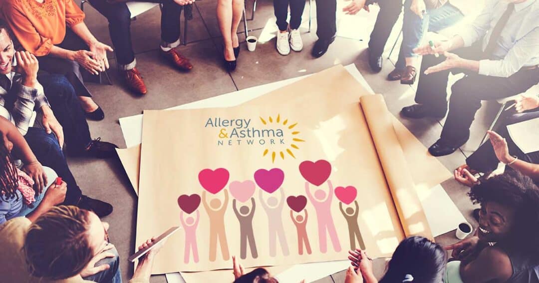 Allergy & Asthma Network 40th Anniversary 2 A diverse group of people sit in a circle around a large poster on the floor. The poster shows an illustration of people holding heart shapes, with the text "Allergy & Asthma Network" and a sun logo.