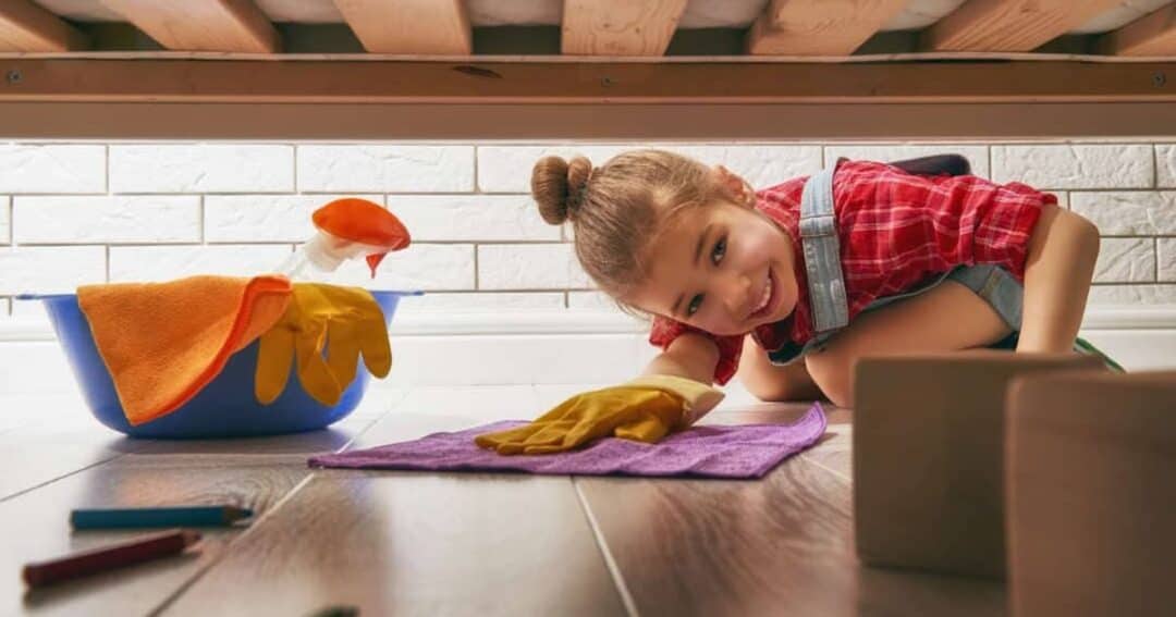 A young girl wearing a red shirt and overalls is smiling while cleaning under a bed. She has a cleaning cloth and yellow gloves. Nearby, a bucket with cleaning supplies is visible on a wooden floor.