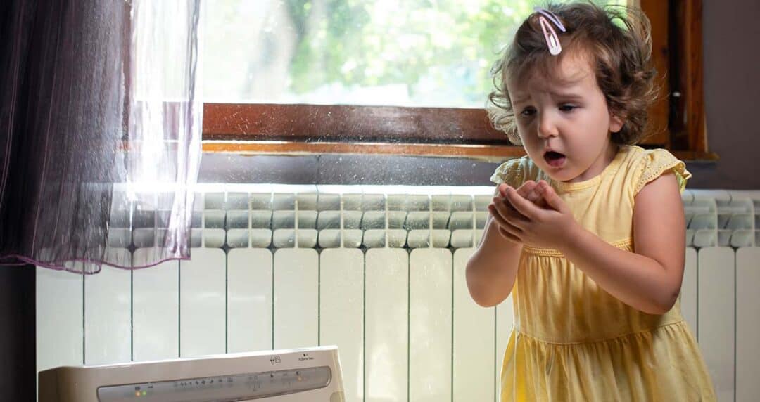 Indoor Air Quality and Indoor Air Pollution 1 A young girl in a yellow dress stands by a radiator and holds her hands to her face as she sneezes, with sunlight streaming through a window, illuminating dust particles in the air.
