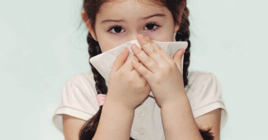 A young girl with braided hair is wearing a white shirt. She is holding a tissue or handkerchief over her nose and mouth, suggesting she might be sneezing or covering her face. The background is a soft, pale color.